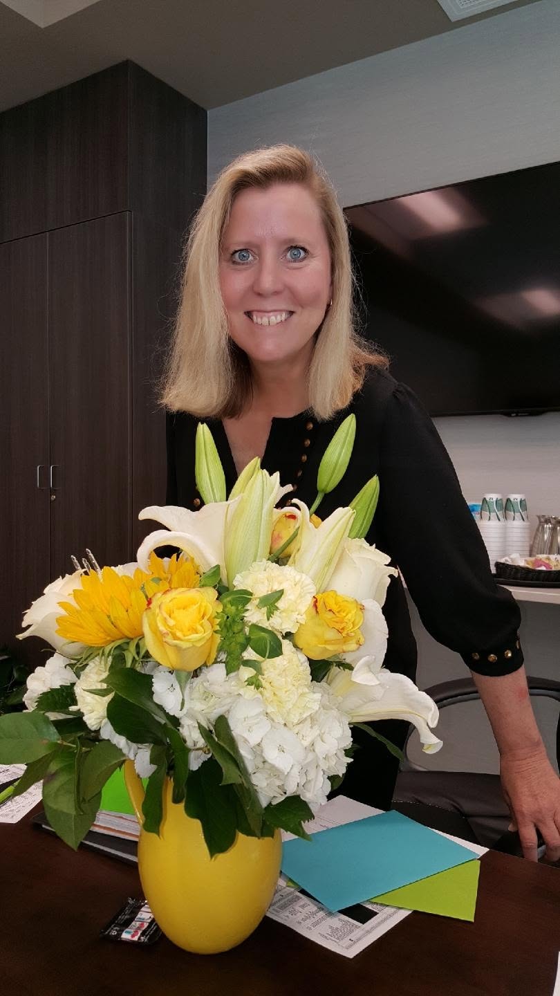 Patti standing behind a vase of yellow and white flowers, smiling.